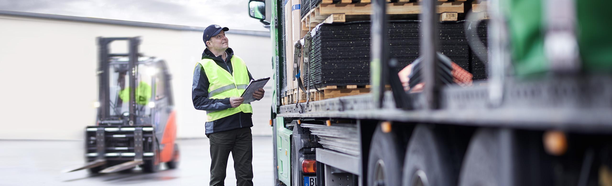Lagerarbeiter mit Warnweste kontrolliert die Verladung mithilfe von Antirutschmatten, im Hintergrund ein Gabelstapler in Bewegung.
